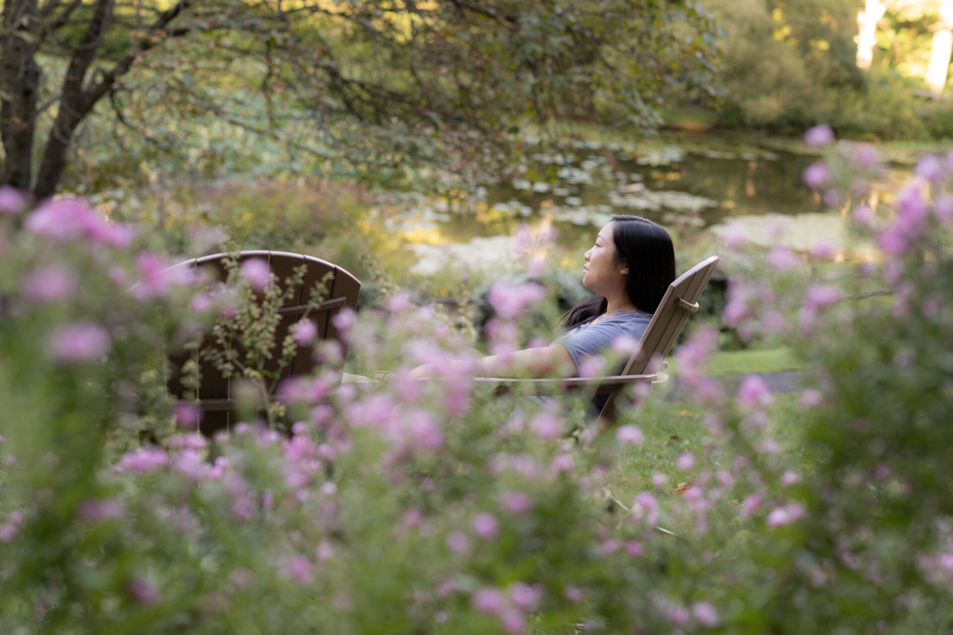 A young woman with dark hair leans back in an Adirondack chair surrounded by pink fluffy aster blooms.