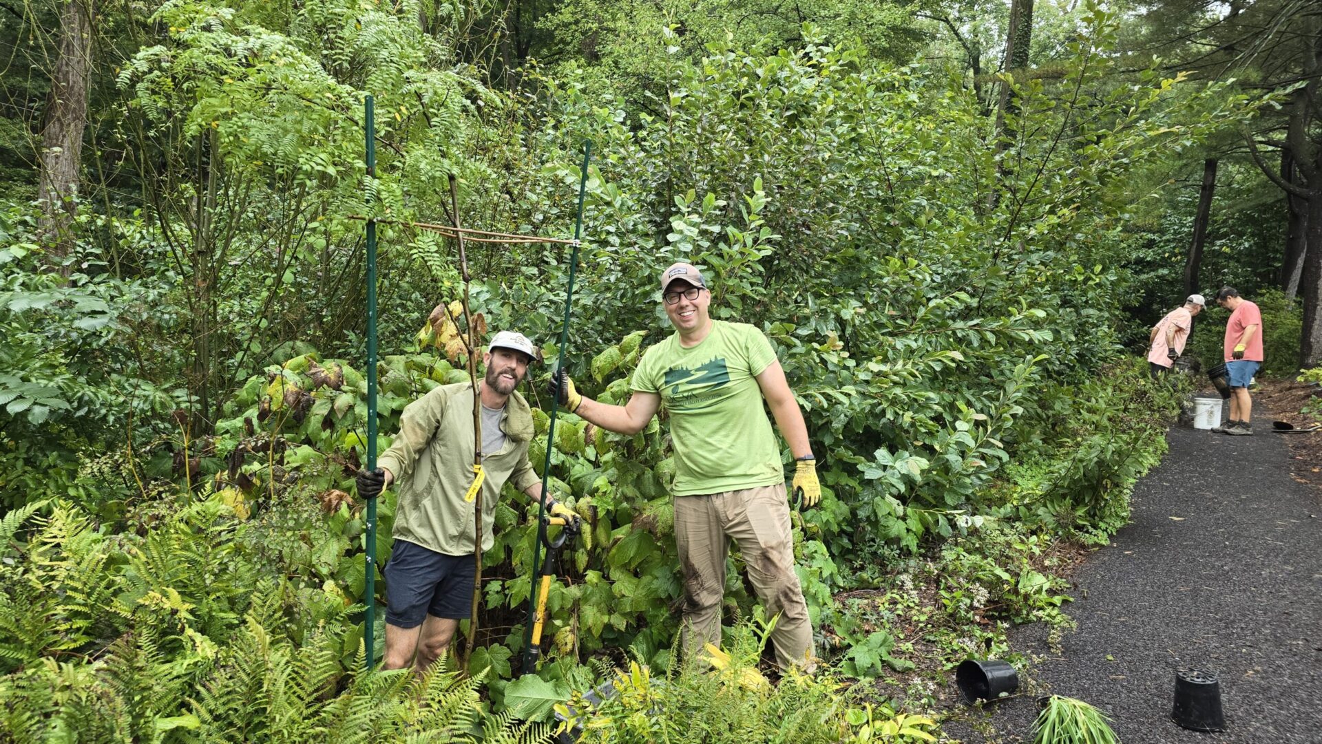 Two men wearing green shirts and baseball caps pose near a small tree during a volunteer planting day.