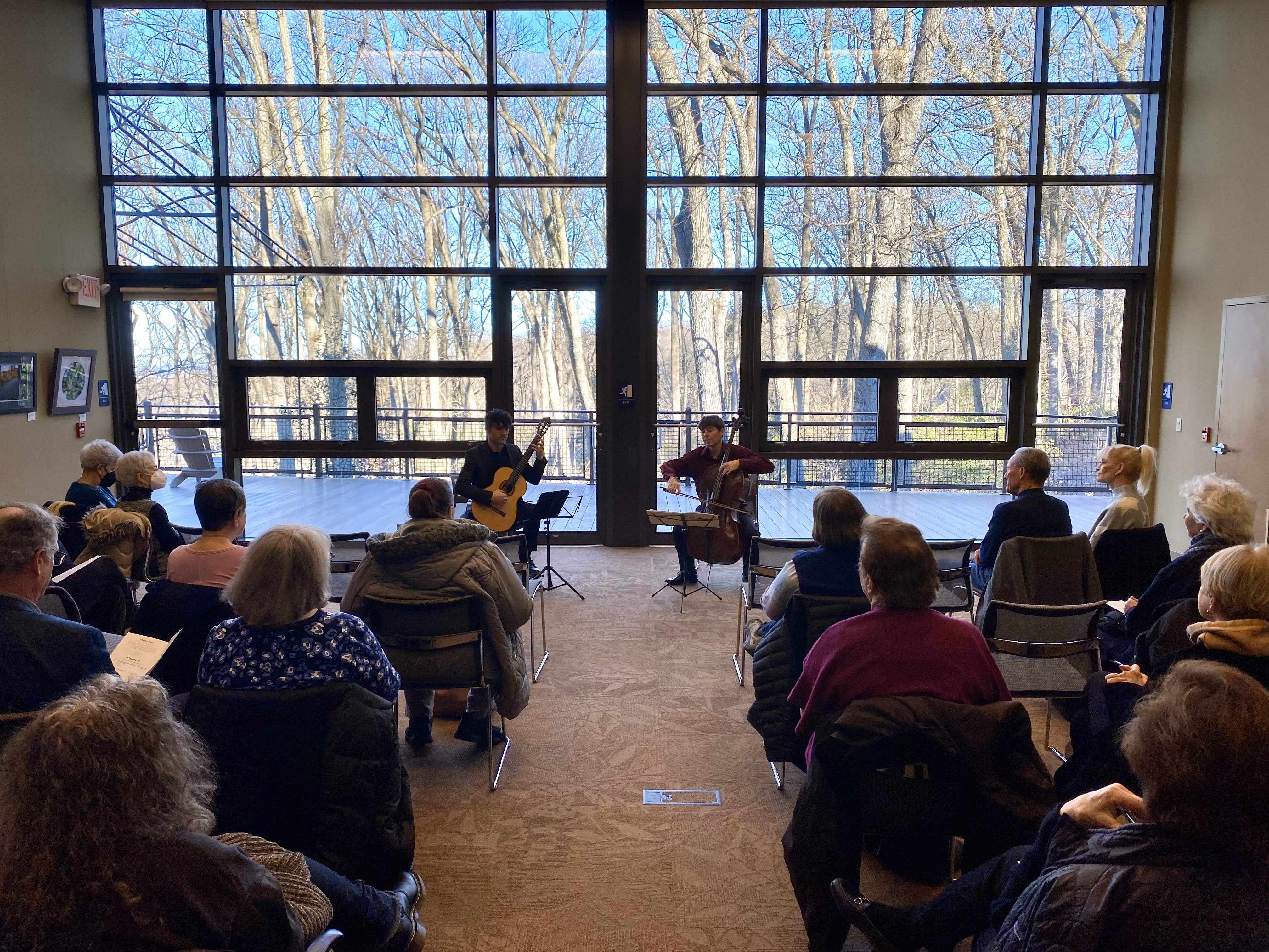 A group of people watch a duo of musicians playing cello and guitar against the backdrop of windows looking out into a wintry garden.