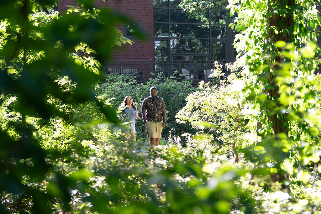 A pair of young adult visitors walk down a sunlit, leafy path with the education center in the background.